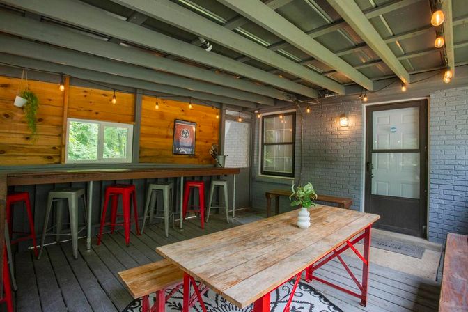 Cozy covered backyard patio with a rustic wood picnic table on red metal legs, matching benches, high bar counter with red and gray metal stools, warm string lights, potted plant centerpiece, and gray brick walls with an exterior door.