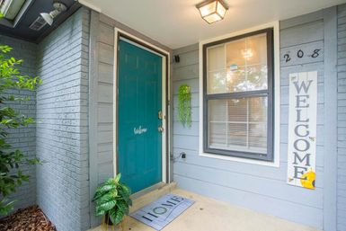 Cozy front porch with a teal entry door reading "Welcome", gray siding, vertical WELCOME sign, "HOME" doormat, potted plants, hanging greenery and a lit porch light