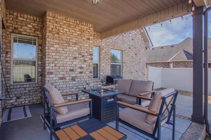 Covered suburban backyard patio with brick exterior, cushioned outdoor seating arranged around a lit fire-pit table, string lights overhead, a grill by the window, and a white privacy fence.