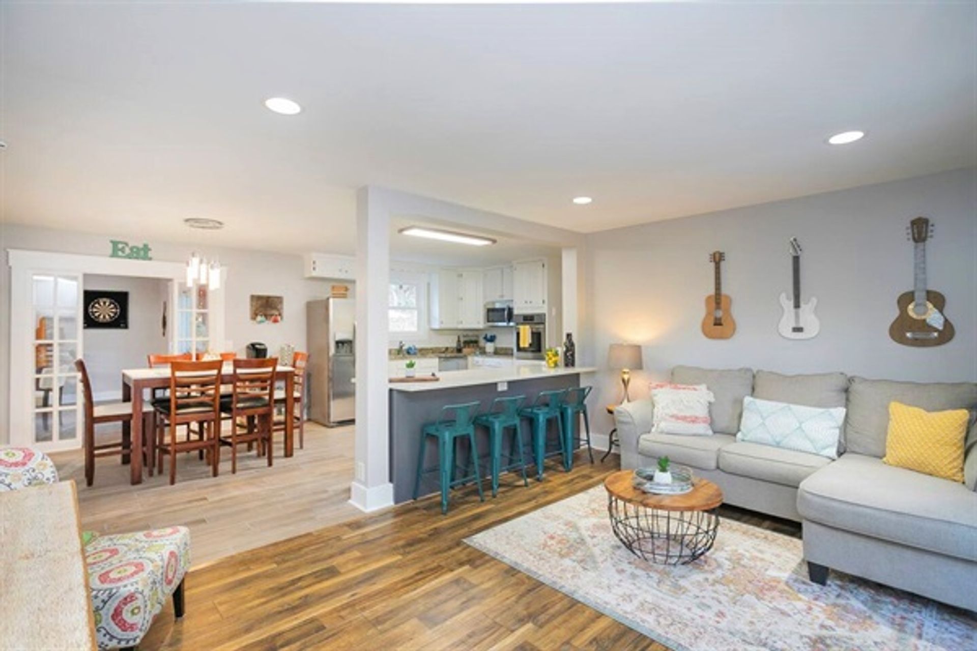 Bright open-concept living room and kitchen with hardwood floors, gray sectional, colorful pillows, three guitars on the wall, teal metal bar stools at a breakfast island, wooden dining table, stainless appliances and a patterned area rug