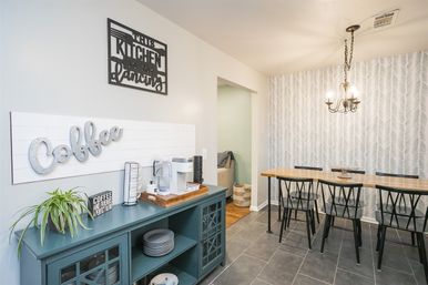 Cozy modern farmhouse kitchen breakfast nook with teal coffee-station cabinet and metal “Coffee” wall art, white coffee maker on a wooden tray, long wooden bar table with black metal chairs under a chandelier, gray patterned accent wall and tile floor.