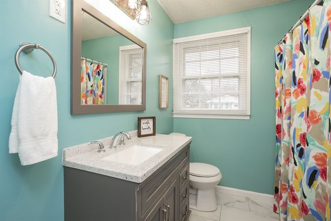 Bright teal bathroom with a gray vanity and speckled white countertop, chrome faucet, white toilet, colorful floral shower curtain, towel ring and window with blinds.
