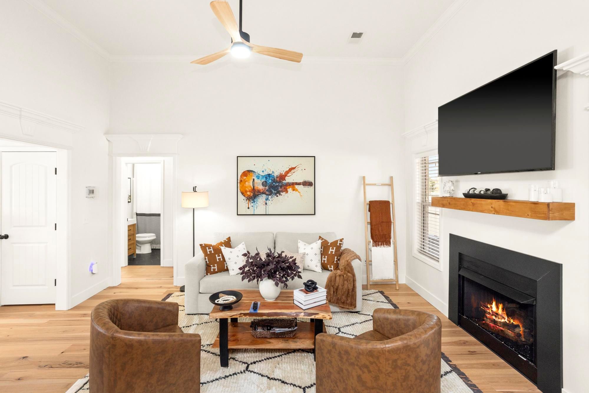 Cozy modern living room with a lit fireplace and wall-mounted flat-screen TV, neutral sofa with patterned pillows, two leather accent chairs, wooden coffee table and hardwood floors under a ceiling fan, colorful guitar artwork on the wall.
