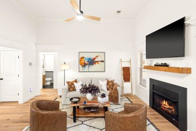 Cozy modern living room with a lit fireplace and wall-mounted flat-screen TV, neutral sofa with patterned pillows, two leather accent chairs, wooden coffee table and hardwood floors under a ceiling fan, colorful guitar artwork on the wall.