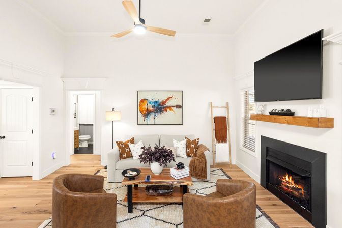 Cozy modern living room with a lit fireplace and wall-mounted flat-screen TV, neutral sofa with patterned pillows, two leather accent chairs, wooden coffee table and hardwood floors under a ceiling fan, colorful guitar artwork on the wall.