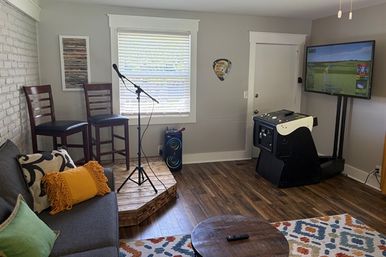 Cozy home entertainment room with a small raised wooden stage, microphone on a stand and two bar stools, gray sofa with colorful pillows, portable speaker, arcade-style golf game with wall-mounted TV showing a golf simulator, hardwood floors and patterned rug.