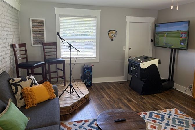Cozy home entertainment room with a small raised wooden stage, microphone on a stand and two bar stools, gray sofa with colorful pillows, portable speaker, arcade-style golf game with wall-mounted TV showing a golf simulator, hardwood floors and patterned rug.