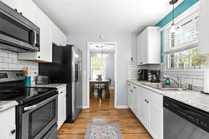 Bright modern galley kitchen with white shaker cabinets, subway tile backsplash, granite-look countertops, stainless steel appliances, wood-look floors and a view into a sunlit dining room.