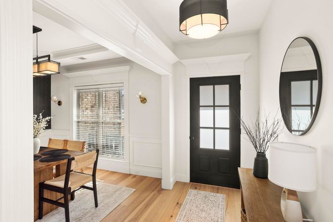 Bright modern entryway and dining area with black frosted-glass front door, oak floors, white trim, round wall mirror, wooden console table, and pendant-lit dining table with woven chairs.