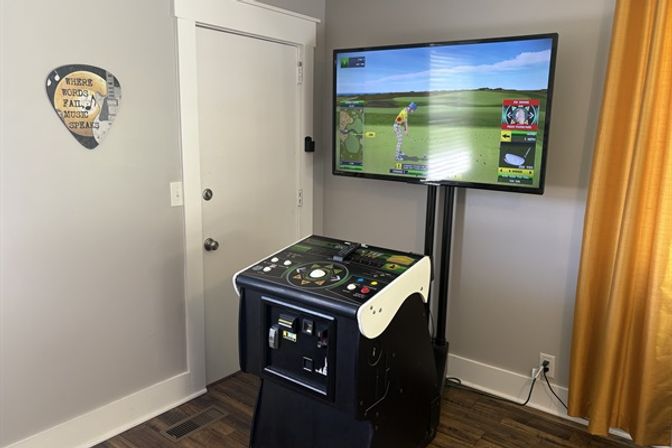 Arcade-style indoor golf simulator in a room corner with a control console and large wall-mounted TV showing a virtual golf course, yellow curtains and door in the background.