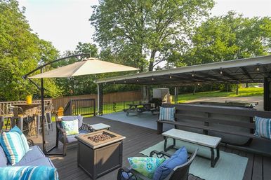 Backyard deck patio outdoor living space with cantilever umbrella, cushioned seating and blue-striped pillows, square fire pit, picnic table under a covered pavilion with string lights, and leafy trees