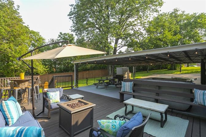 Backyard deck patio outdoor living space with cantilever umbrella, cushioned seating and blue-striped pillows, square fire pit, picnic table under a covered pavilion with string lights, and leafy trees