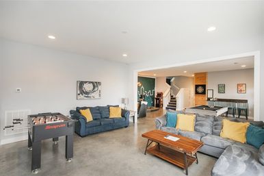 Bright modern finished basement recreation room with foosball and pool tables, dartboard, comfy sectional sofas with yellow pillows, wooden coffee table, and open staircase.