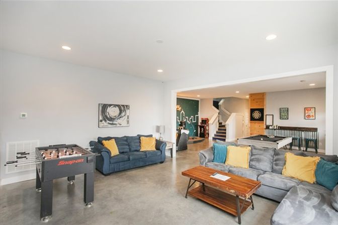 Bright modern finished basement recreation room with foosball and pool tables, dartboard, comfy sectional sofas with yellow pillows, wooden coffee table, and open staircase.