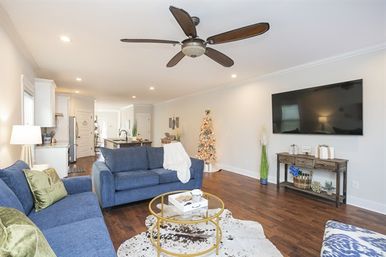Open-concept modern living room with blue sofas, round glass coffee table on cowhide rug, wall-mounted flat-screen TV, ceiling fan, hardwood floors, and a small decorated Christmas tree by the kitchen island.