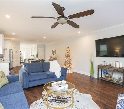 Open-concept modern living room with blue sofas, round glass coffee table on cowhide rug, wall-mounted flat-screen TV, ceiling fan, hardwood floors, and a small decorated Christmas tree by the kitchen island.