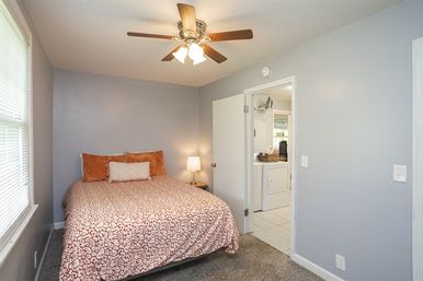 Cozy guest bedroom with patterned coral duvet, orange throw pillows, neutral gray walls, ceiling fan and bedside lamp, window with blinds, and an open door revealing an adjoining laundry area with washer/dryer.