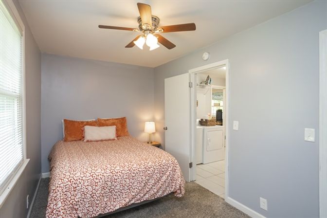 Cozy guest bedroom with patterned coral duvet, orange throw pillows, neutral gray walls, ceiling fan and bedside lamp, window with blinds, and an open door revealing an adjoining laundry area with washer/dryer.