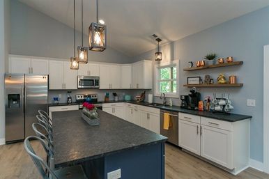 Bright modern open kitchen with a large dark island and metal bar stools, white cabinets, stainless-steel appliances, pendant lights, floating wood shelves and hardwood floors.