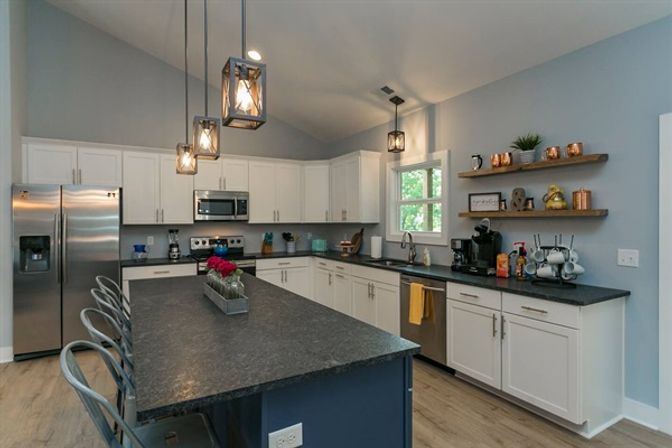 Bright modern open kitchen with a large dark island and metal bar stools, white cabinets, stainless-steel appliances, pendant lights, floating wood shelves and hardwood floors.