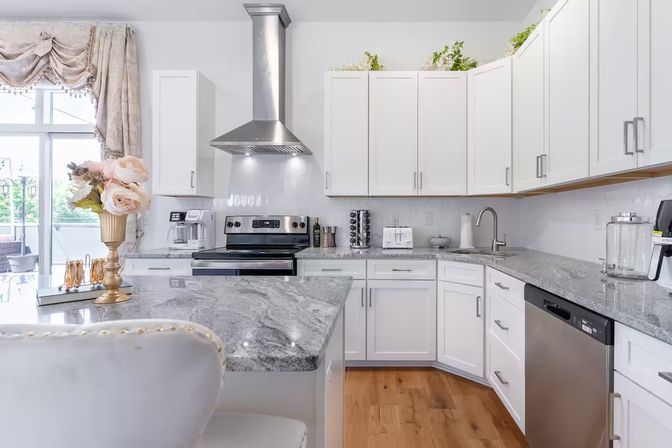 Sunlit modern white kitchen with gray marble island, stainless-steel range and dishwasher, white shaker cabinets, hardwood floors, tufted island seating, and a gold vase of pale roses by a patio door.