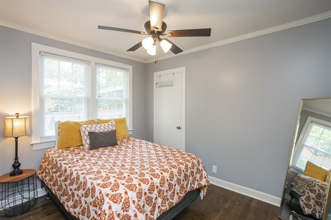 Cozy, light-filled modern bedroom with gray walls, hardwood floors, ceiling fan, double windows, patterned bedspread, bedside lamp, and a leaning full-length mirror.