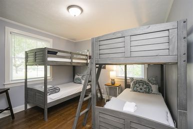 Bright cozy bedroom with two gray wooden bunk beds, white linens and patterned pillows, hardwood floors, bedside table with lamp, and a window letting in natural light.