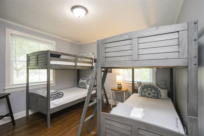 Bright cozy bedroom with two gray wooden bunk beds, white linens and patterned pillows, hardwood floors, bedside table with lamp, and a window letting in natural light.
