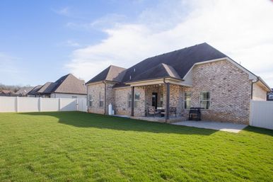 Sunny suburban backyard with lush green lawn, brick ranch-style house, covered patio with seating, and white privacy fence.