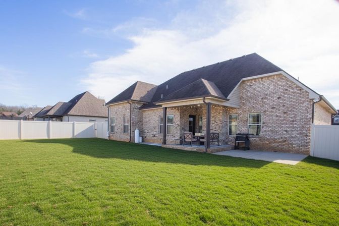 Sunny suburban backyard with lush green lawn, brick ranch-style house, covered patio with seating, and white privacy fence.