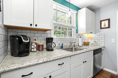 Sunlit modern white kitchen with subway-tile backsplash, gray granite countertop, double stainless sink and dishwasher, and a coffee station with single-serve and drip brewers under a window with white blinds.