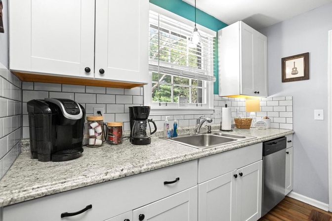 Sunlit modern white kitchen with subway-tile backsplash, gray granite countertop, double stainless sink and dishwasher, and a coffee station with single-serve and drip brewers under a window with white blinds.