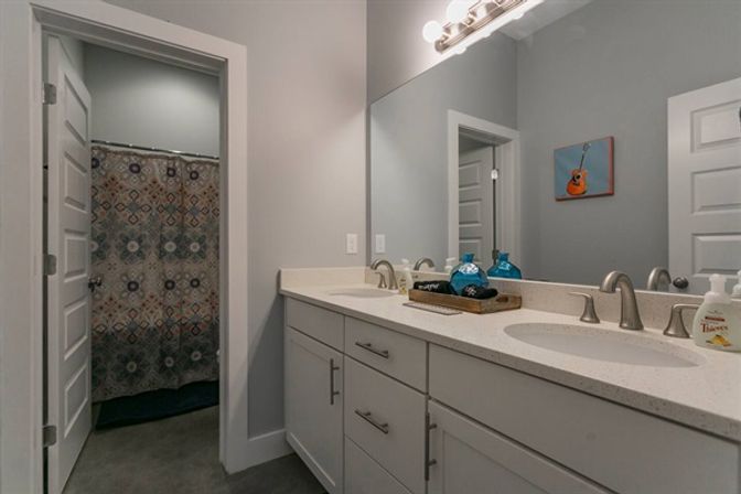 Modern gray bathroom interior with double-sink vanity and quartz countertop, brushed-nickel faucets, large mirror, patterned shower curtain and blue decorative bottles.