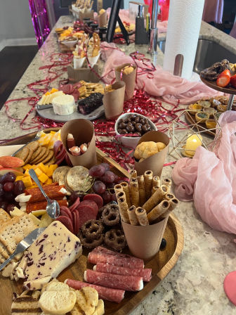 Party-ready charcuterie spread on a marble kitchen island: wooden boards with cheeses (including cranberry cheddar and soft rounds), cured meats, crackers, grapes, chocolate-covered pretzels, wafer rolls and small bites, styled with pink tulle, metallic streamers and a candle for a festive appetizer display.