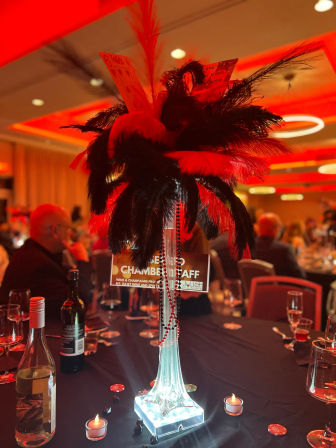 Tall illuminated glass centerpiece topped with red and black feathers, playing cards and red bead strands on a banquet table in a hotel ballroom, surrounded by wine bottles, tealight candles and blurred guests