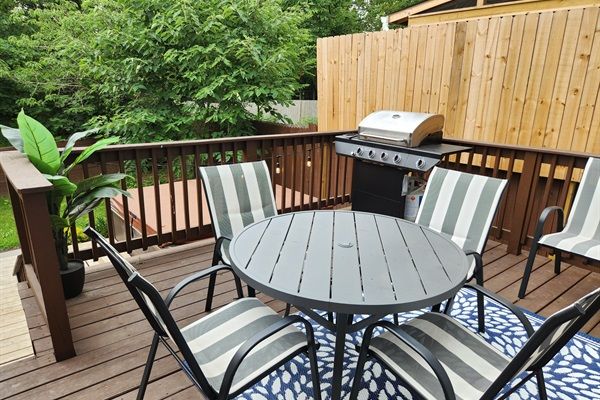 Sunny backyard wooden deck with round gray metal table, four gray-and-white striped patio chairs on a blue rug, propane gas grill, potted plant and wooden privacy fence with trees beyond.