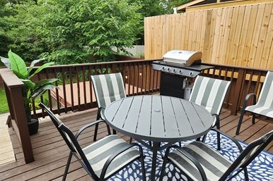 Sunny backyard wooden deck with round gray metal table, four gray-and-white striped patio chairs on a blue rug, propane gas grill, potted plant and wooden privacy fence with trees beyond.