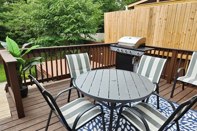 Sunny backyard wooden deck with round gray metal table, four gray-and-white striped patio chairs on a blue rug, propane gas grill, potted plant and wooden privacy fence with trees beyond.