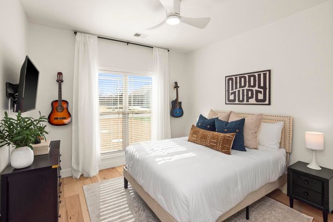 Bright, modern bedroom interior with white walls and wood floors, cozy bed layered with decorative pillows, two acoustic guitars mounted on the wall, wall-mounted TV, potted plant on a dresser, bedside lamp, and a large sunlit window with white curtains.
