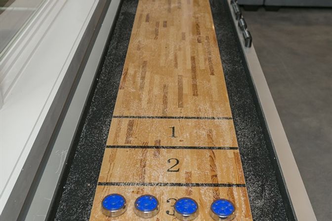 Close-up of an indoor wooden shuffleboard table with numbered scoring zones 1–3 and four blue pucks lined up at the 3-point area