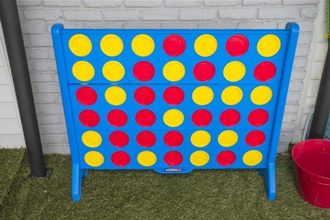 Playful oversized blue four-in-a-row game board with red and yellow discs set against a white brick wall on artificial turf, outdoor patio scene.