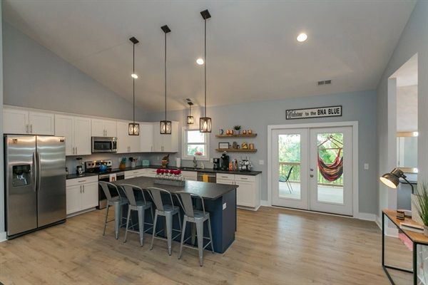 Bright open-concept modern kitchen with a large navy island, five metal bar stools, stainless steel appliances, pendant lights, light wood floors, and French doors to a porch with a colorful hammock.