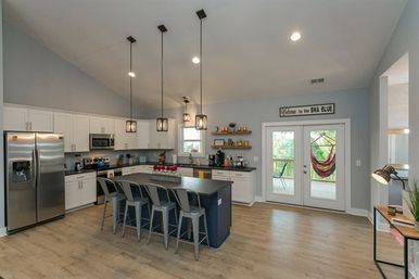 Bright open-concept modern kitchen with a large navy island, five metal bar stools, stainless steel appliances, pendant lights, light wood floors, and French doors to a porch with a colorful hammock.