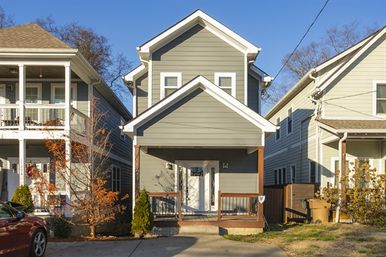 Cozy two-story gray siding house with gabled roof and covered front porch on a narrow suburban street, flanked by neighboring homes, small front trees and clear blue sky.