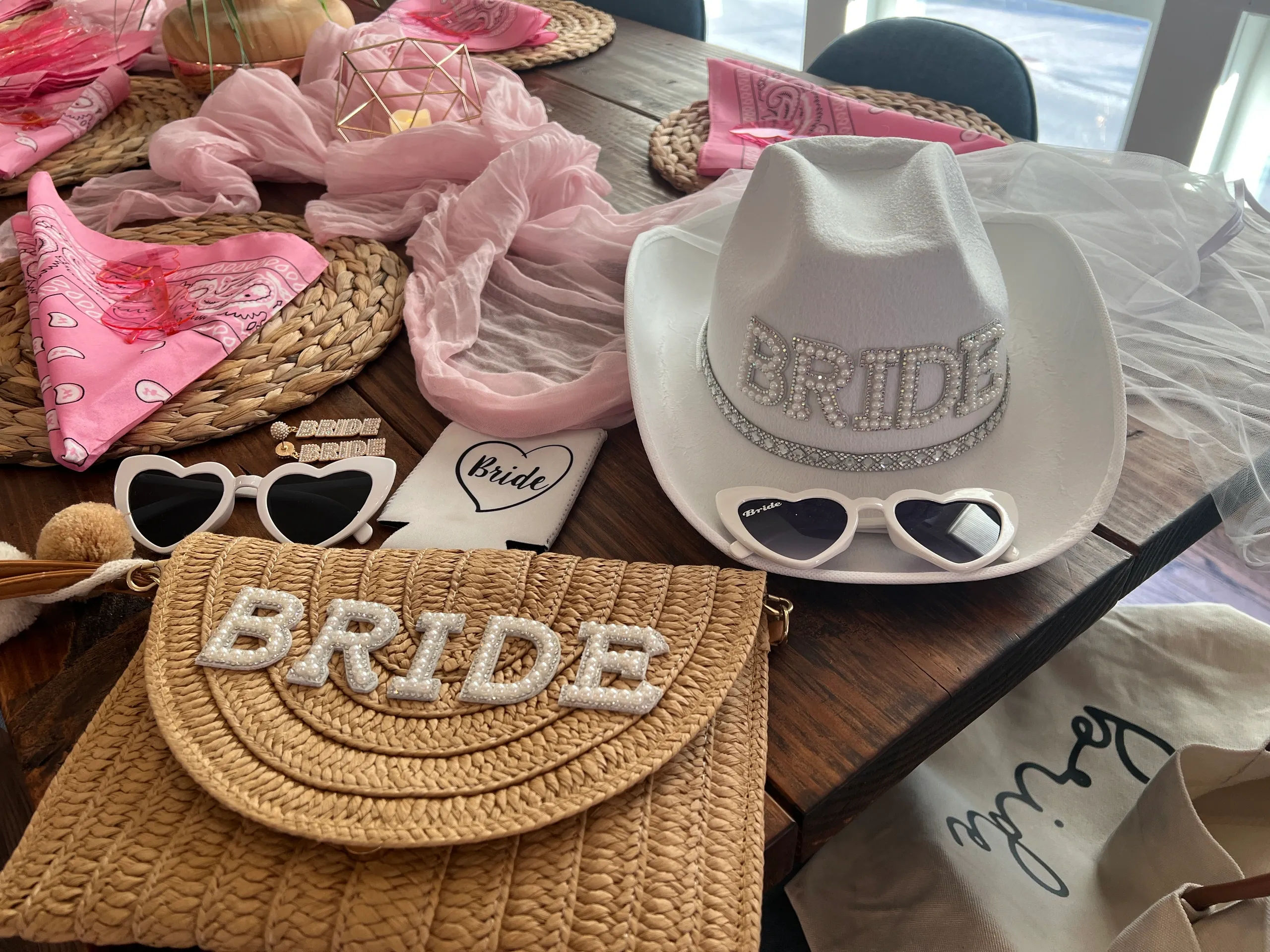 Bachelorette party bride accessories on a wooden table: rhinestone BRIDE white cowboy hat, straw BRIDE purse, heart-shaped sunglasses, pink bandanas and veil.