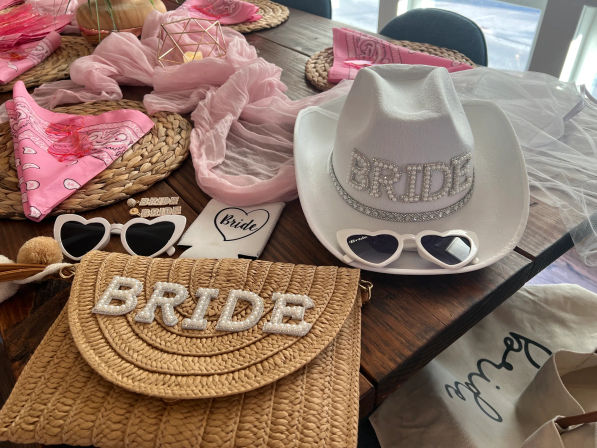 Bachelorette party bride accessories on a wooden table: rhinestone BRIDE white cowboy hat, straw BRIDE purse, heart-shaped sunglasses, pink bandanas and veil.