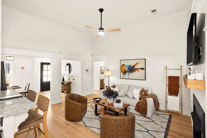 Cozy bright open-plan living room with white walls and high ceiling, light hardwood floors, gray sofa with patterned cushions, two brown leather chairs, wood coffee table on a geometric rug, colorful abstract guitar art, ladder blanket rack, black ceiling fan, and a granite kitchen island with wooden bar stools.