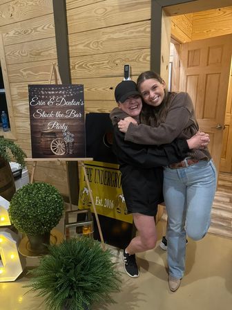 Two smiling friends hugging inside a wood‑paneled venue next to a rustic “Stock the Bar” party sign, potted greenery and warm event lighting.