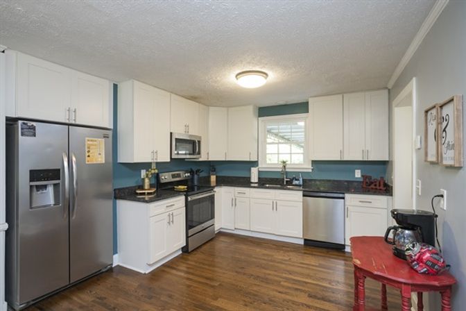 Bright, cozy kitchen with white shaker cabinets, stainless steel fridge, oven, microwave and dishwasher, dark granite countertops, teal backsplash, hardwood floors, window above sink and a red side table with a coffee maker.