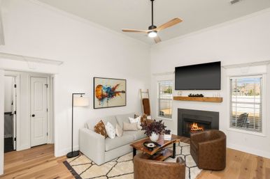 Bright cozy modern living room in a suburban home with hardwood floors, light-gray sofa, leather armchairs, wood coffee table, gas fireplace beneath a wall-mounted TV, abstract guitar artwork and large windows.